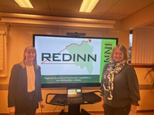 To damer foran et stort tv-skjerm med logo til forskningsprosjektet Redinn